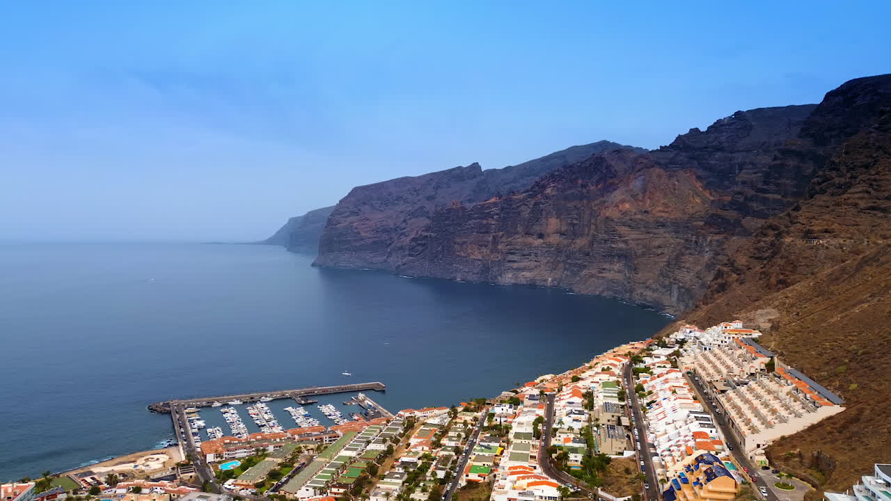 Flight above the residential area on the slope of the bare rock. Approaching the yacht club at the coast of the Atlantic Ocean. Aerial perspective on Tenerife Island, Spain