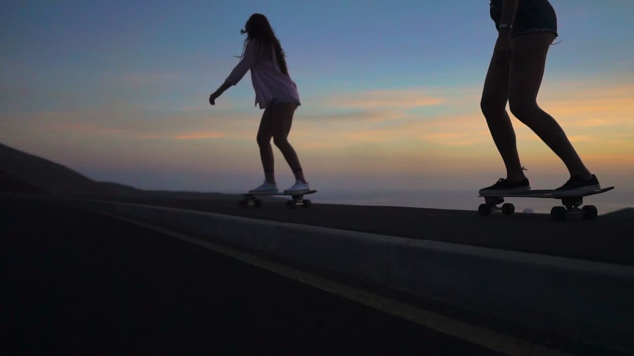 Slow-motion footage showcases two friends in shorts skateboarding on a road during sunset. The mountains and a picturesque sky form an alluring backdrop