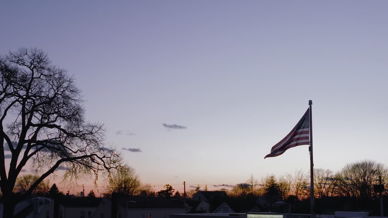 Sebuah bidikan udara, pada ketinggian rendah, bendera Amerika berkibar tertiup angin saat matahari terbenam, dalam gerakan lambat dengan siluet pepohonan di latar belakang.
