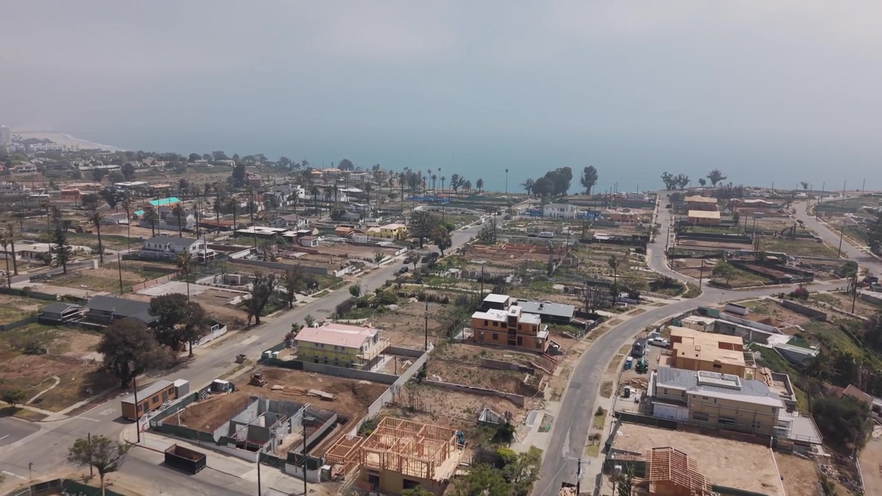 Le conseguenze dell'incendio di Pacific Palisades che mostrano lotti vuoti e fondamenta bruciate, con la ricostruzione in corso accanto a case sopravvissute e palme sotto un cielo velato. California, USA. Sorvolo aereo.