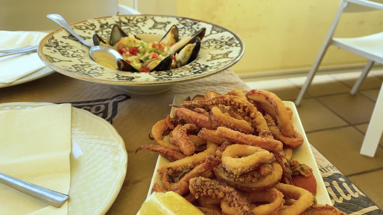 Slow-motion close-up of traditional Cantabrian tapas: fried calamari rings with lemon and mussels in vinaigrette. Warm tones reflect the cozy atmosphere of a seaside restaurant terrace