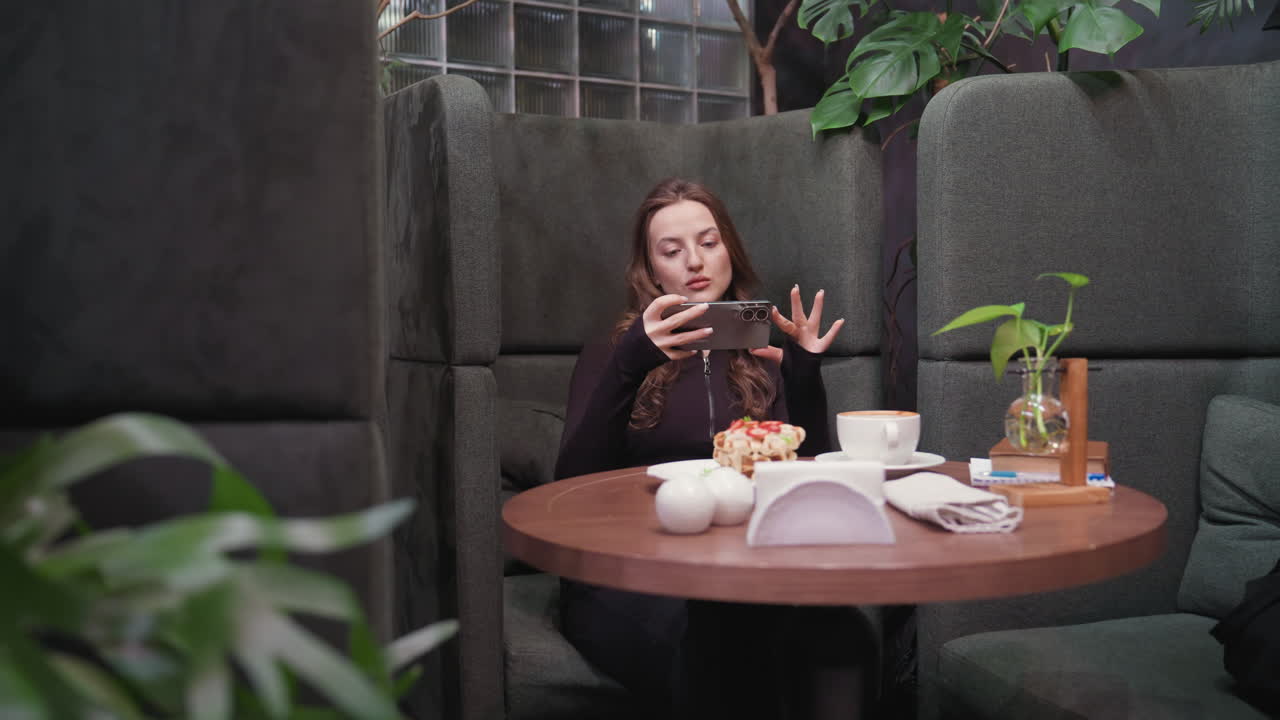 Young lady seated indoors taking picture of waffles and coffee on round wooden table decorated with small plant and napkins, surrounded by cushioned booths and leafy green ambiance