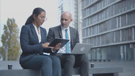 business colleagues with laptop and tablet talking and working on project together while sitting at office building outside