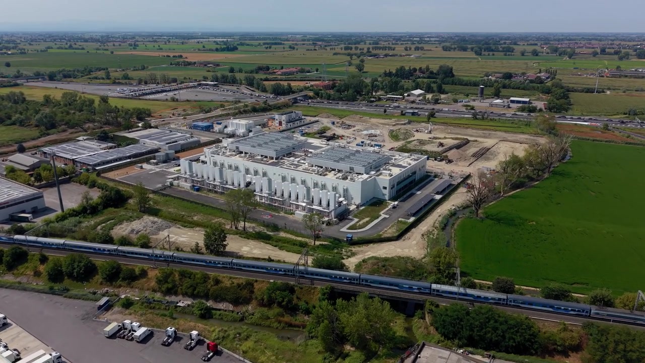 Avvicinamento aereo con drone verso un moderno data center mentre un treno passeggeri si muove lungo la ferrovia adiacente. La scena collega infrastrutture digitali, reti di trasporto e sviluppo industriale moderno.