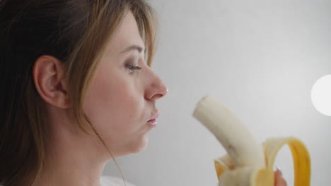 close up of woman eating banana under bright vanity light indoors showcasing healthy morning snack routine with soft facial expression and gentle lighting in relaxed personal grooming environment
