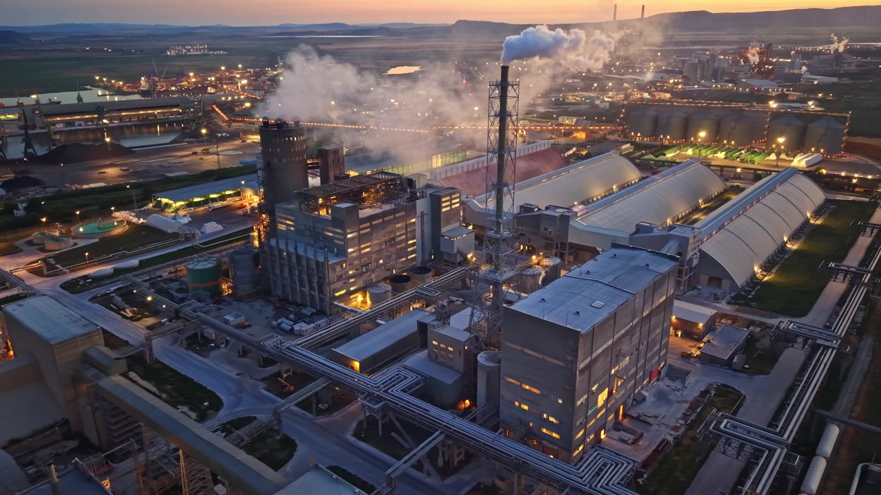 Industrial site during sunset with smoke rising from factories and lights turning on around the complex
