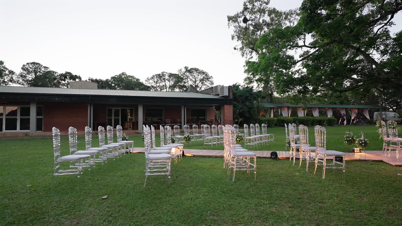 Allestimento per cerimonia di matrimonio all'aperto con corridoio decorato, composizioni floreali ed eleganti sedie trasparenti.