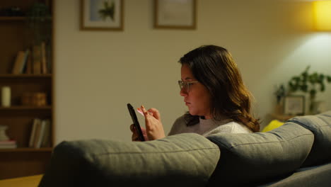 young woman spending evening at home sitting on sofa with mobile phone scrolling through internet or social media