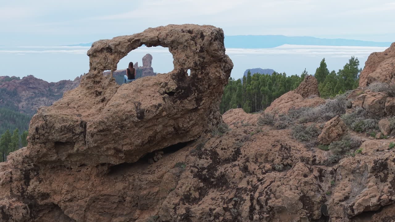 Cinematic aerial shot of a woman sitting in the Window of Roque Nublo (Gañifa), with Roque Nublo majestically in the background. Gran Canaria, Spain.