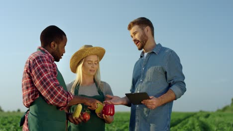 Young-Pretty-Woman-And-Two-Multiethnic-Men-Farmers-Standing-In-The-Green-Field-With-Harvested-Vegetables-In-Hands-And-One-Man-With-A-Tablet-Device