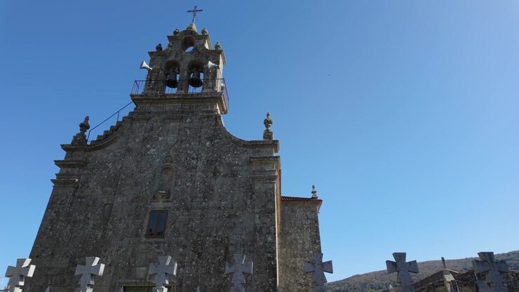 Vista dal basso della chiesa di Santa María A Real de Porqueira ad A Forxa, Ourense, che evidenzia la sua storica facciata in pietra, il campanile a vela ornato e l'architettura barocca sotto un cielo azzurro limpido