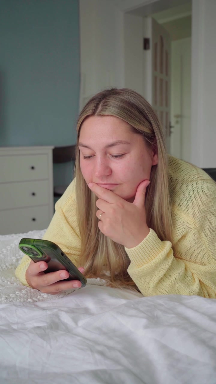 Avslappet ung kvinne i gul genser ligger på sengen på et lyst soverom, smilende mens hun leser gode nyheter på smarttelefonen sin