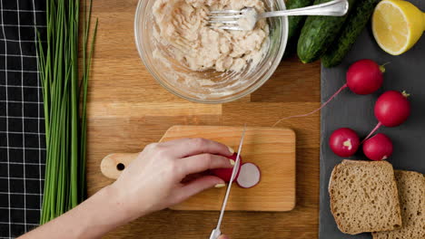 Arriba-Hacia-Abajo-De-Recoger-Un-Rábano,-Rebanarlo-Y-Cortarlo-En-Trozos-Pequeños-Para-Preparar-Un-Sándwich-Vegetal-Saludable-Esparcido-En-Una-Mesa-De-Cocina-De-Madera-Colorida,-4k-25fps