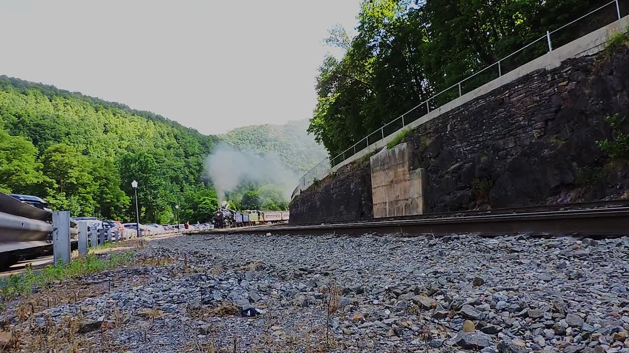 A steam train navigates through lush green mountains, releasing puffs of white smoke into the clear blue sky. The idyllic summer landscape creates a perfect backdrop for this journey.