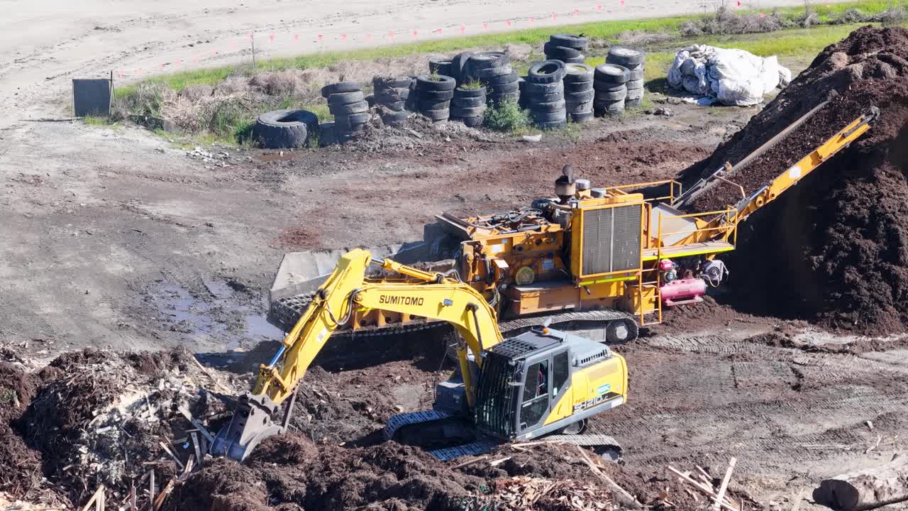 Drone footage captures an excavator loading mulch into a processing machine in a sunlit industrial landscape