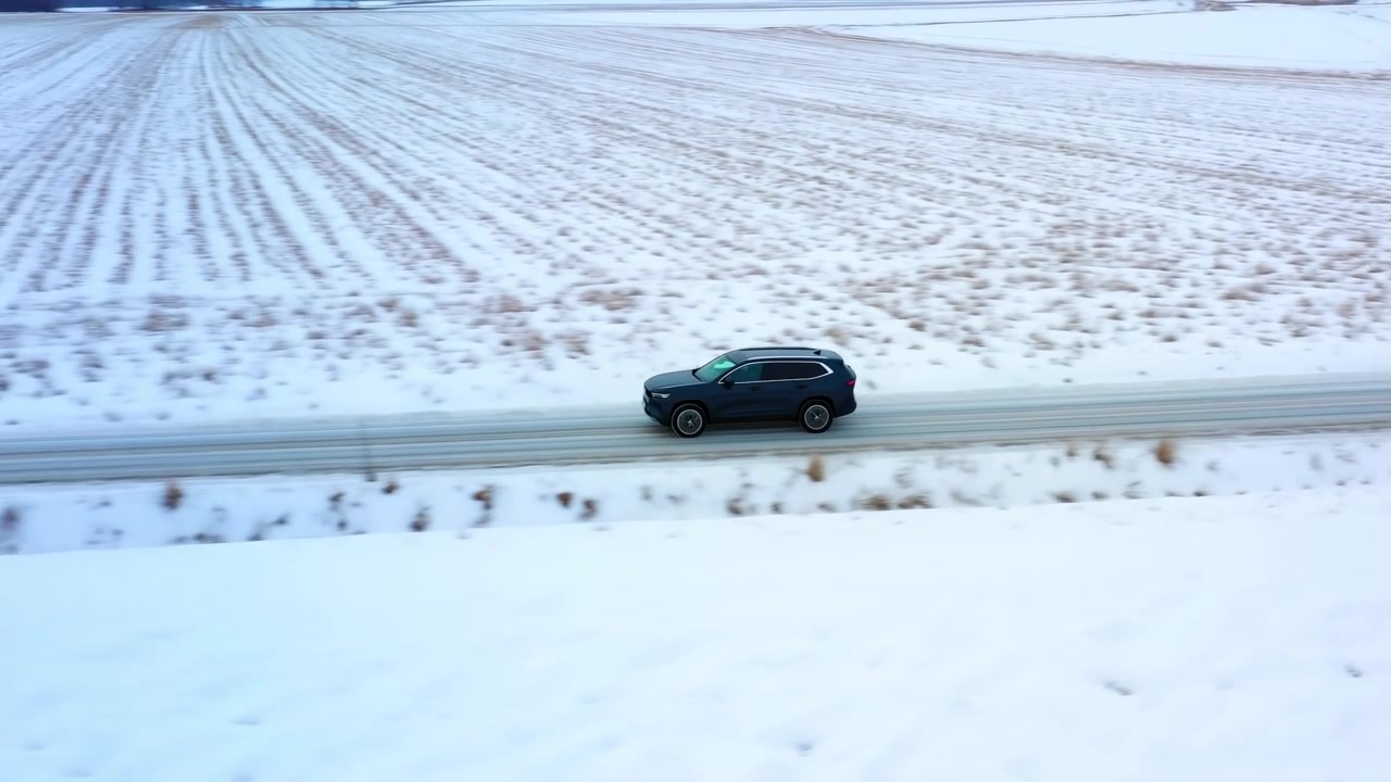 Un SUV blu scuro si muove lungo una stretta strada rurale delimitata da campi agricoli invernali pianeggianti, con chiazze di neve, stoppie marroni, terreno ghiacciato, luce diurna soffusa e ampi dintorni vuoti, con SUV, auto, veicolo, guida.