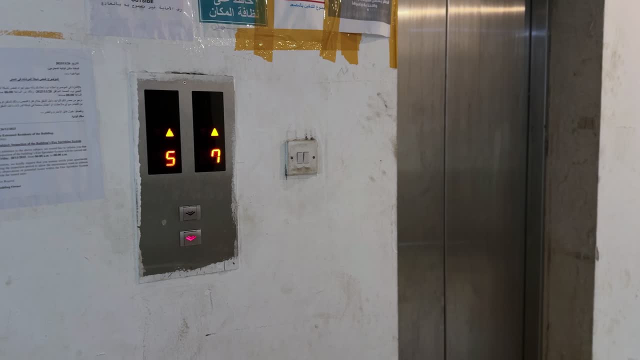 Primer plano de un panel de ascensor interior de edificio que muestra números de piso rojos mientras una mano presiona un botón de bajada iluminado bajo luz artificial.