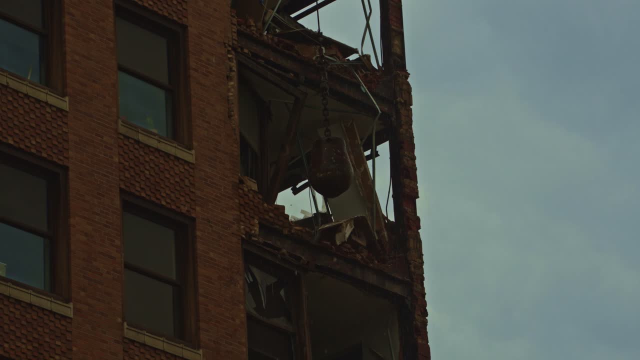 Close-up shot of a wrecking ball smashing through the upper floors of a partially demolished brick high-rise during a downtown demolition project.