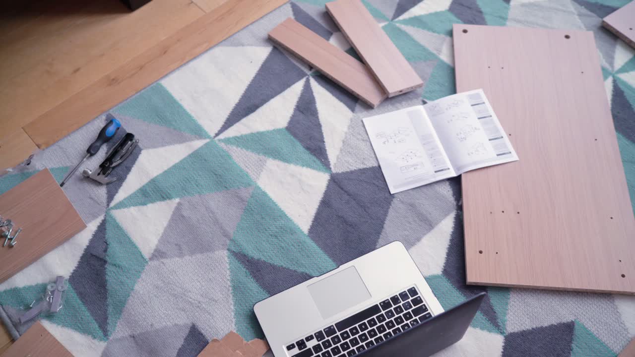 Messy disassembled furniture parts on the ground carpet inside the house with bolts and screws scattered around with a laptop