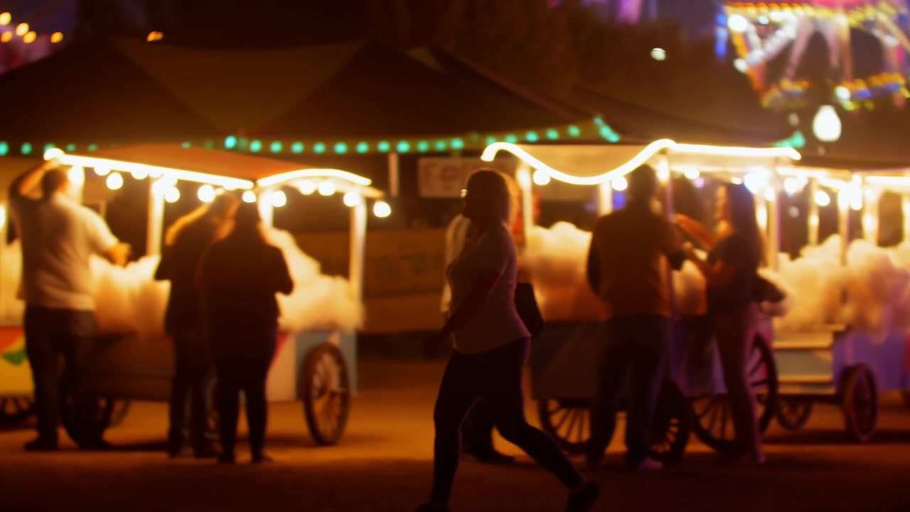 Silhuet af en fodgænger bevæger sig forbi glødende candyfloss-vogne ved siden af farverige tivolilys på et natligt karneval, med afslappet gang, nærliggende menneskemængde og festlig stemning.