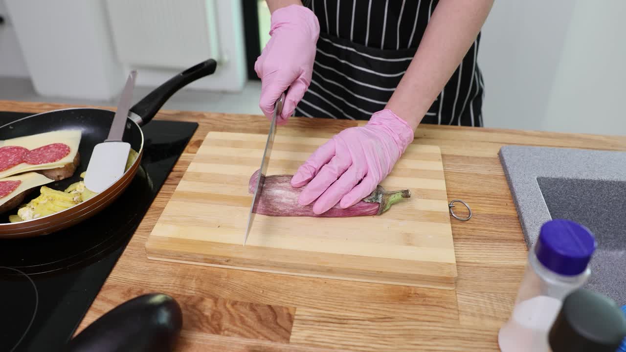 Person wearing pink gloves cutting an eggplant on a wooden board in a kitchen