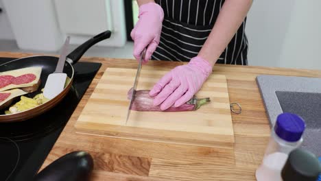 person wearing pink gloves cutting an eggplant on a wooden board in a kitchen