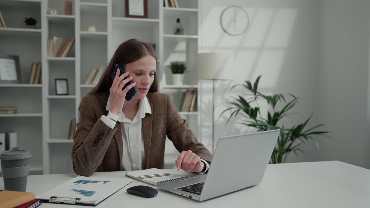 Kvinde på kontoret, der taler i telefon, mens hun bruger bærbar computer