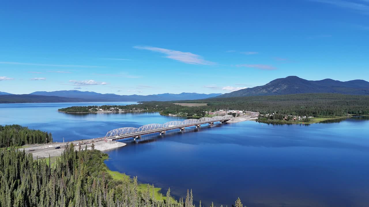 An aerial perspective captures a majestic bridge spanning a vast, tranquil blue lake, flanked by lush green forests and distant, hazy mountains under a clear, sunny sky in Canada