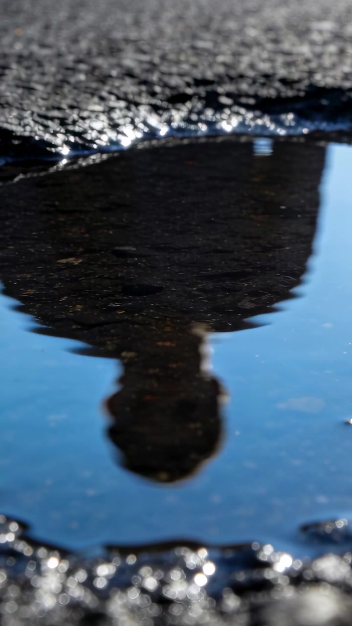 Vertical video: Walking reflected man in dark coat making ripples in puddle on wet asphalt, sun