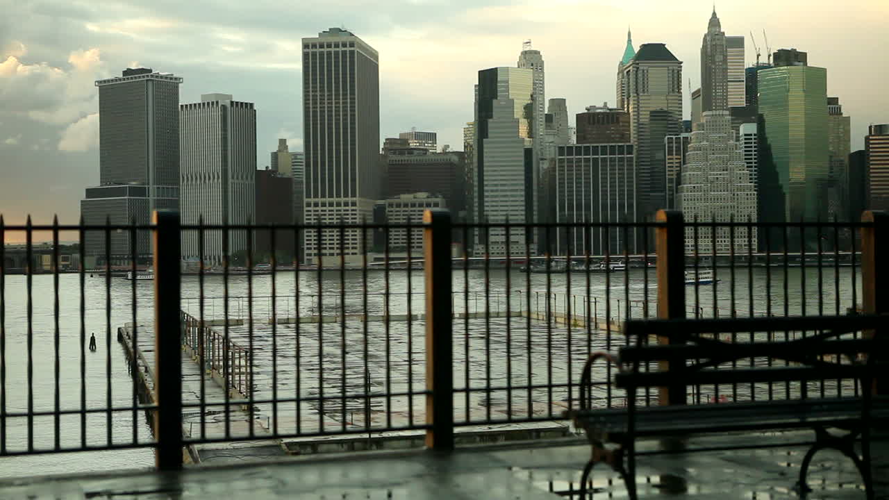 Dolly shot of New York City skyline across the East River from Brooklyn with a park bench in the foreground.