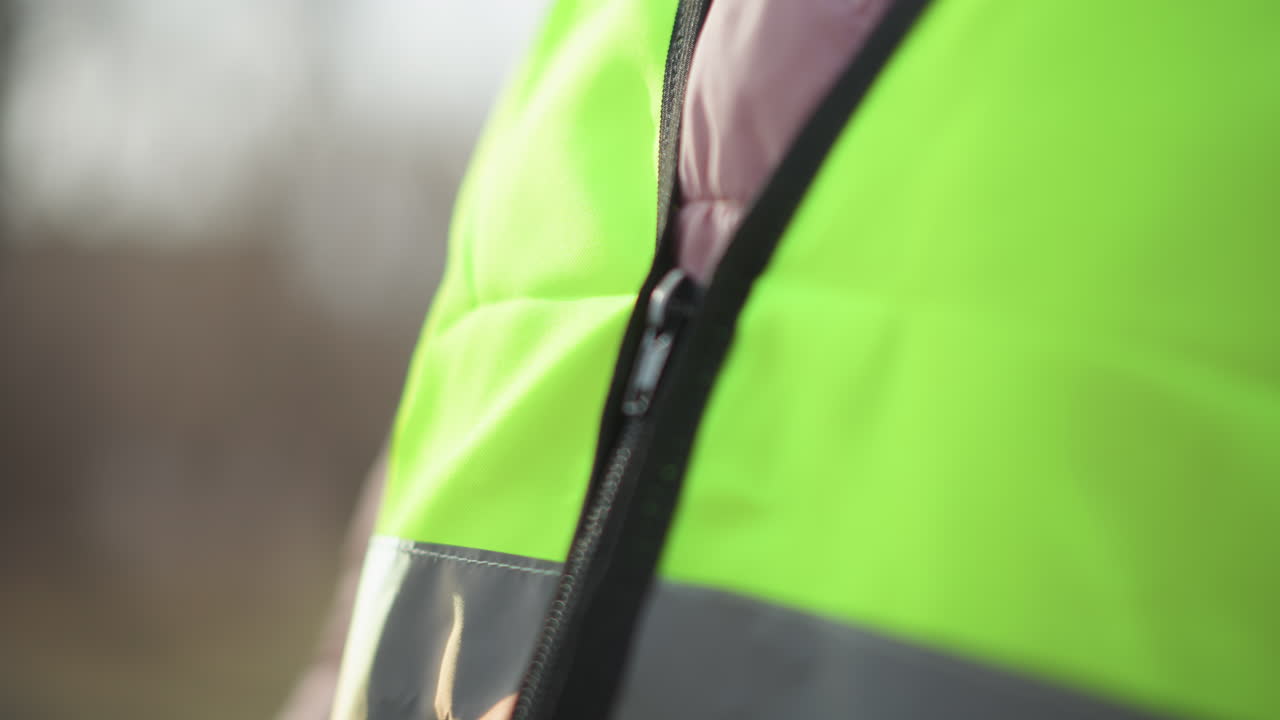 Close-up blurred view of person in pink puffy jacket zipping bright reflective safety vest outdoors during daytime, hands pulling zipper, neon material with grey reflective stripe partially in focus