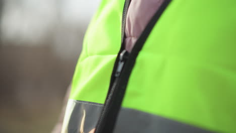 close-up blurred view of person in pink puffy jacket zipping bright reflective safety vest outdoors during daytime, hands pulling zipper, neon material with grey reflective stripe partially in focus