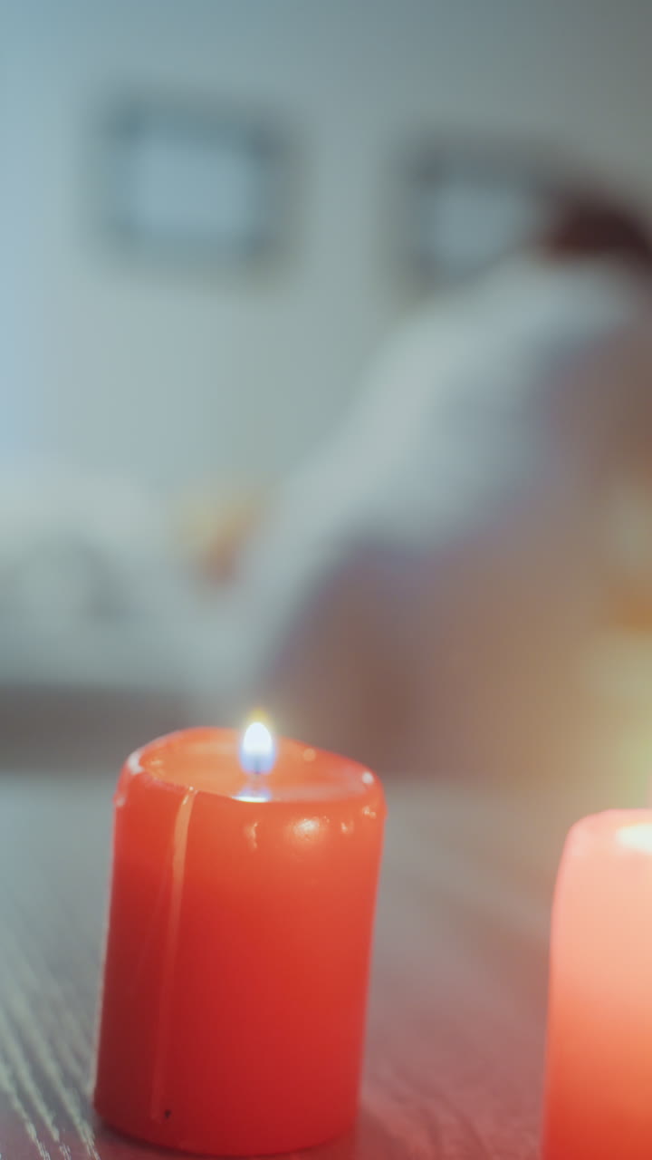 Lilin merah di latar depan adegan pijat di rumah, praktisi membungkuk di atas klien di ranjang, latar belakang fokus lembut, cahaya hangat, unit microcurrent di dekatnya, rutinitas kesehatan malam yang intim dengan aroma menenangkan