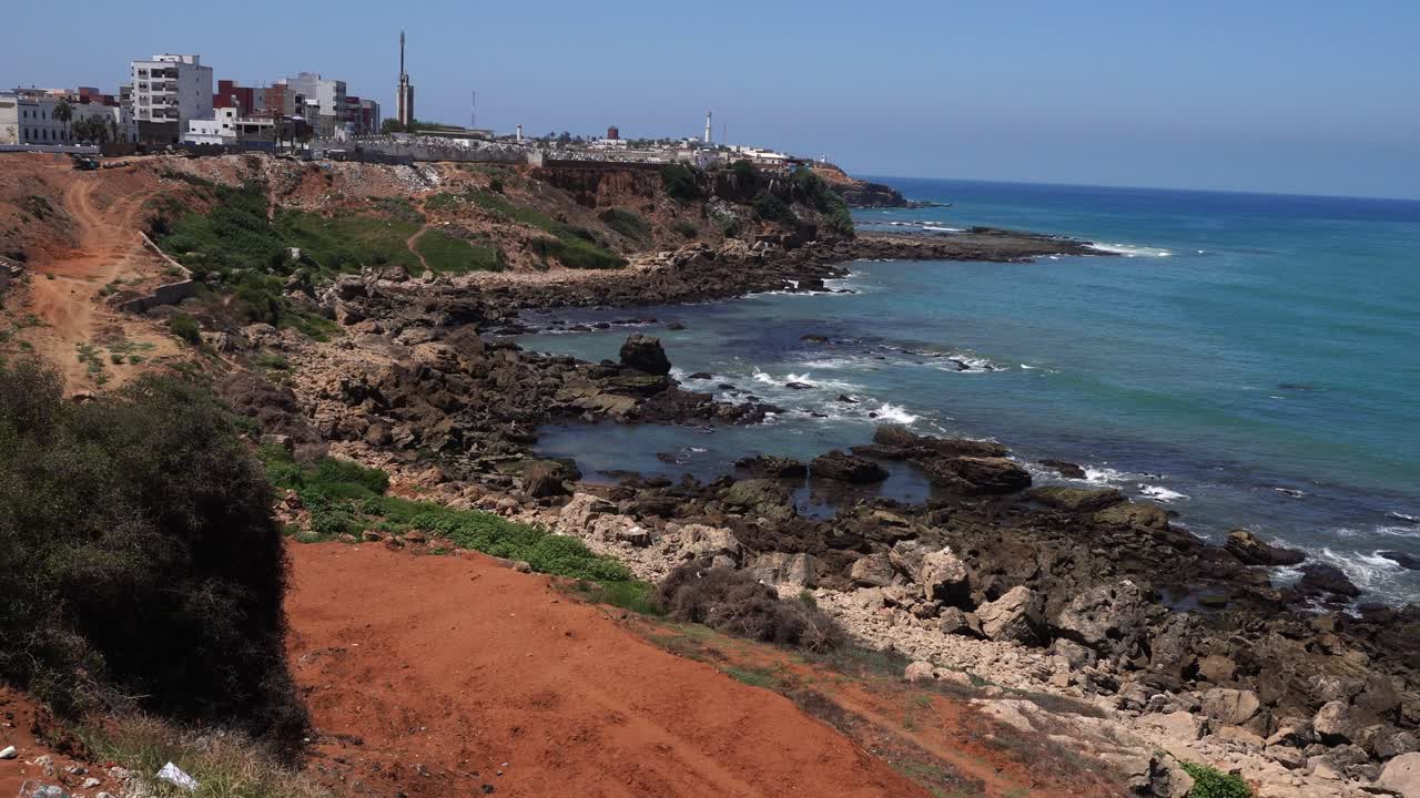 An amazing cluster of incredible rocks stands on the seashore, with waves gently hitting them and a few structures beneath an incredible blue sky