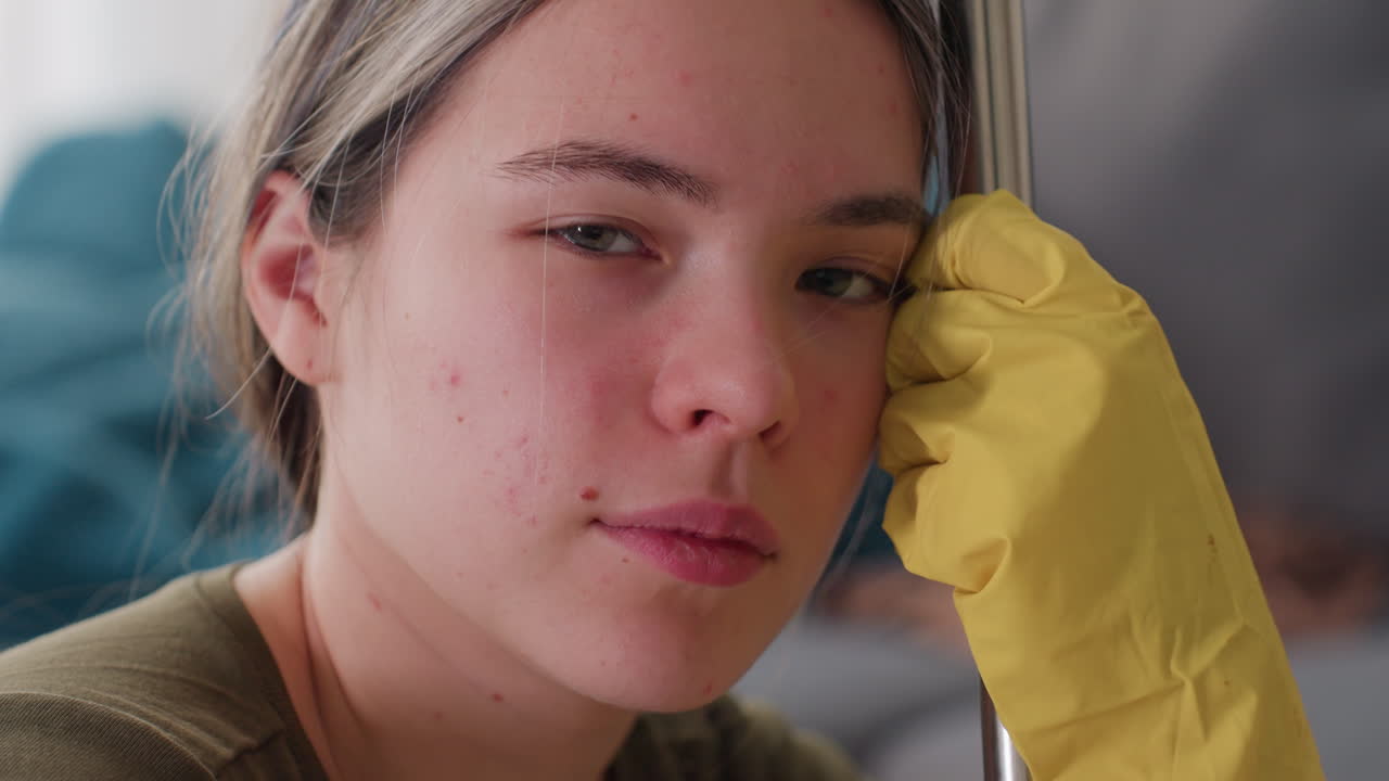 Young woman wearing yellow gloves seated resting head against mop stick looking calm and reflective with close up of face in messy living room background