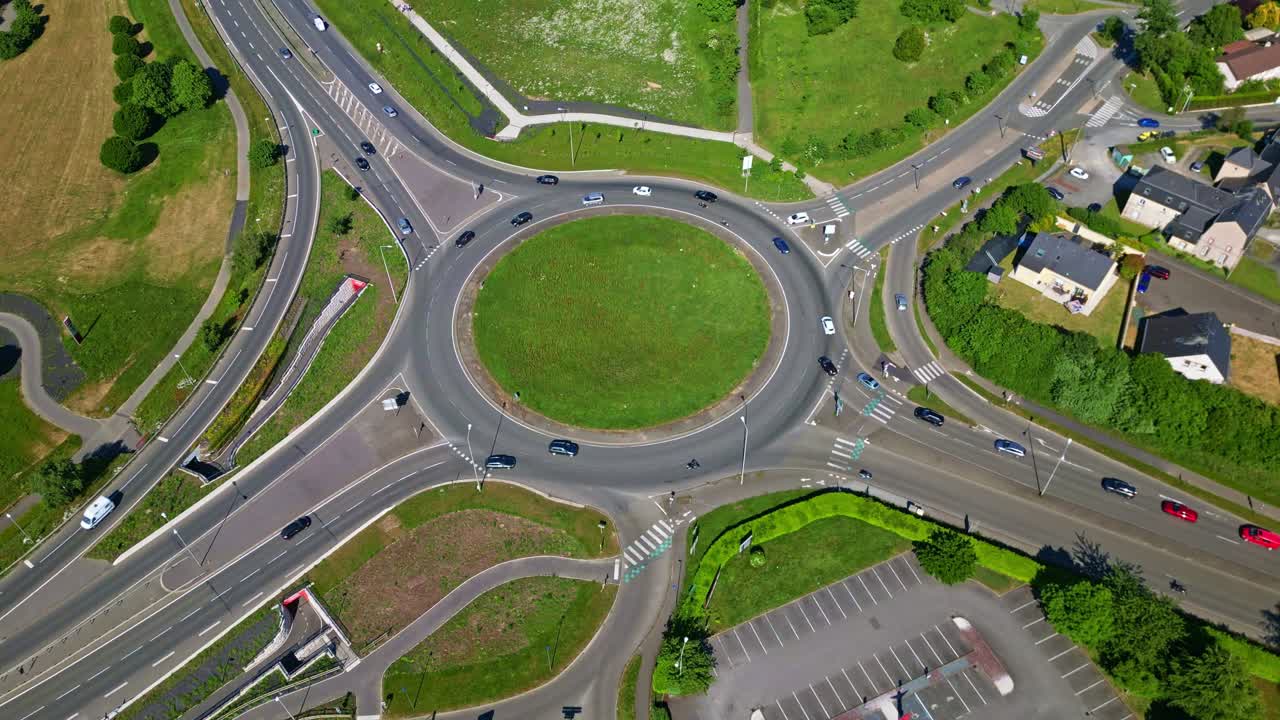 Drone top-down view ascends from roundabout revealing parking, houses, fields, cars, grass, and sunny weather - France
