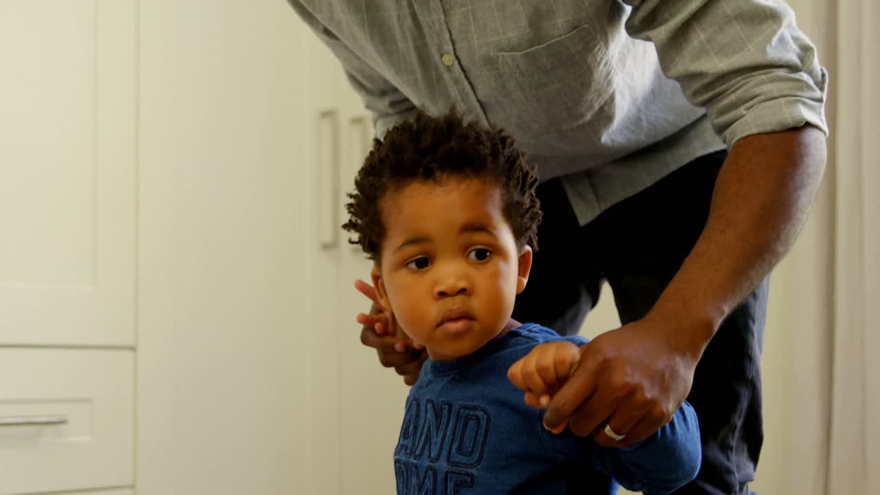 Front view of young black father teaching his son to walk in a comfortable home 4k