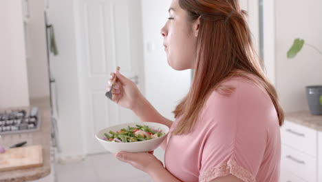 happy plus size biracial woman eating vegetable salad in kitchen, slow motion