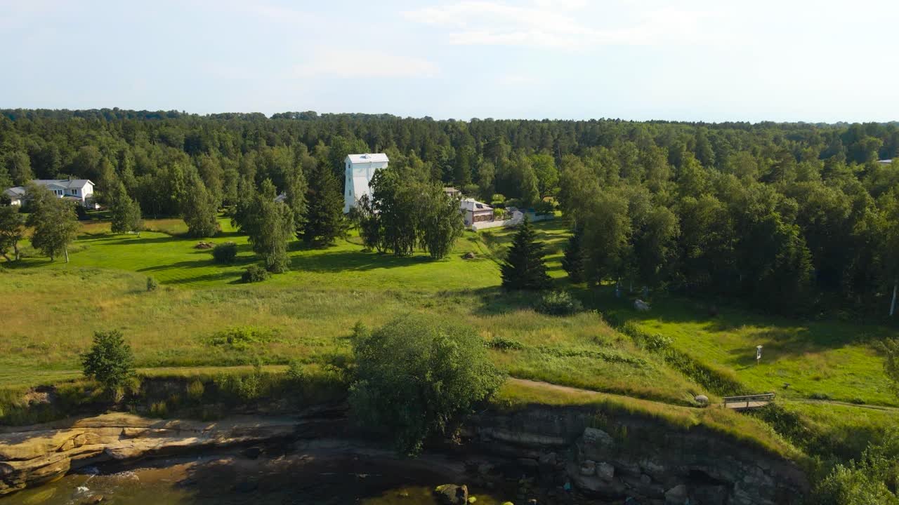 Aerial drone footage flying higher above brown and green Baltic ocean sea water during summer and revealing a large and tall wooden white old lighthouse on a cliff bank shore in Muraste or Suurupi