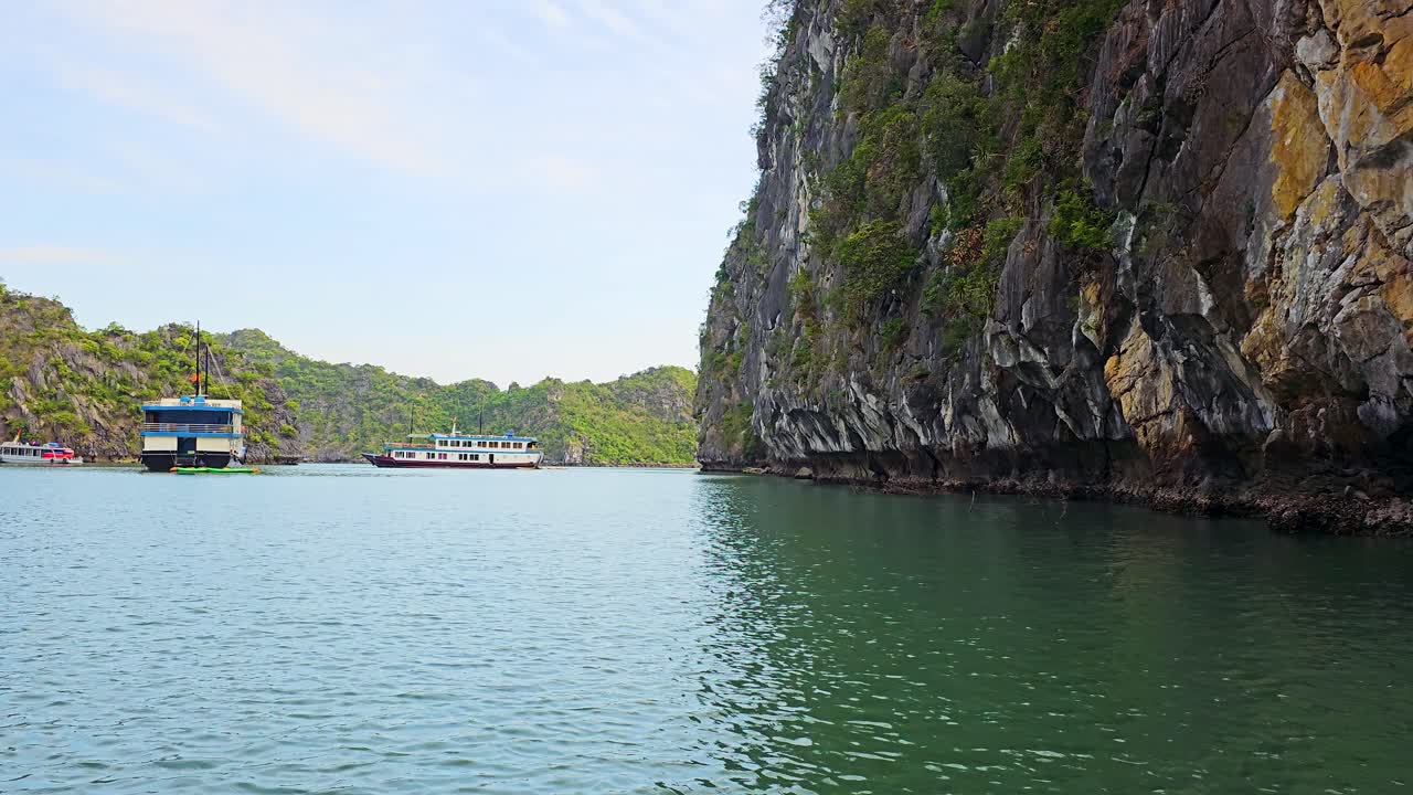 Limestone karst cliffs tower over emerald waters in Vietnam's Ha Long Bay, where traditional cruise boats navigate through the tranquil seascape dotted with floating structures against a serene sky