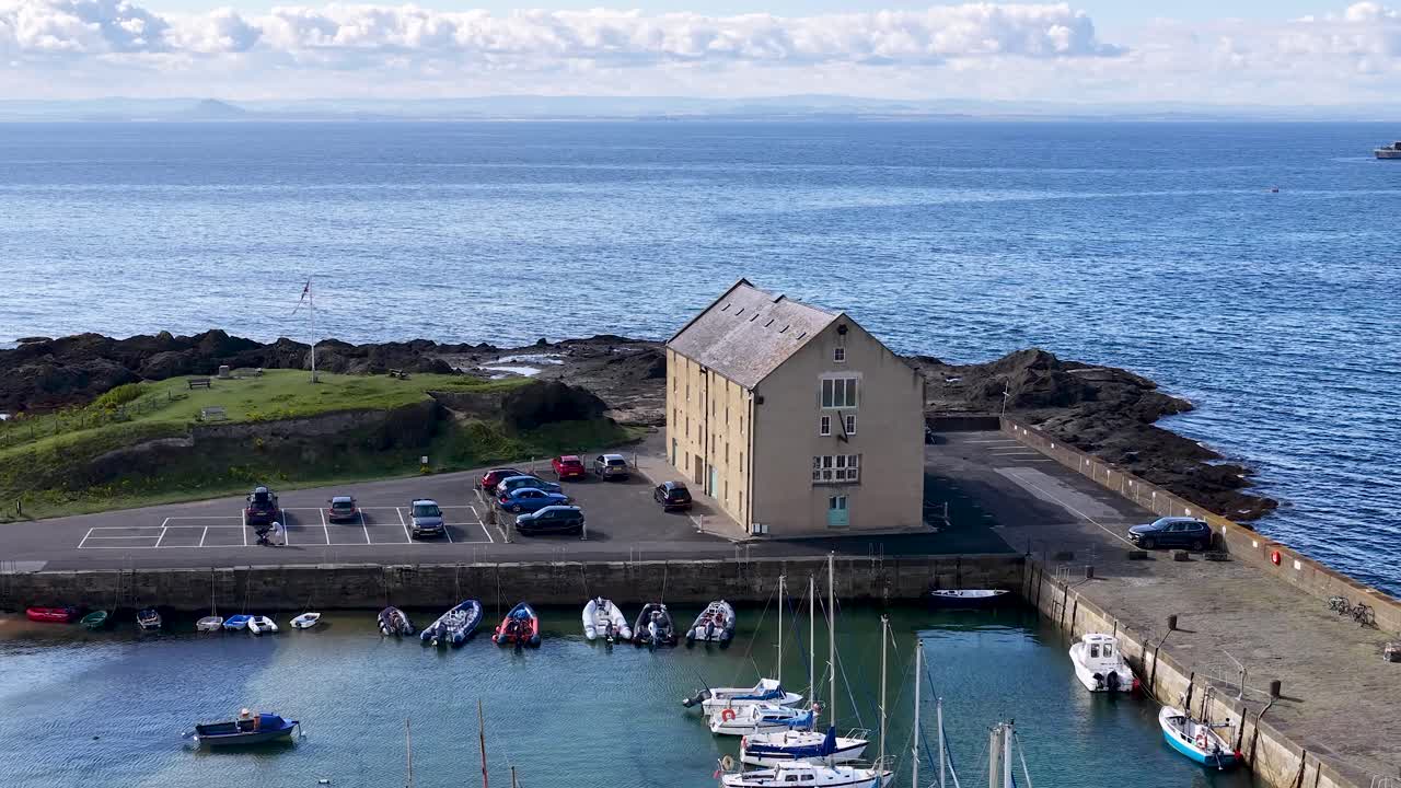 Drone glides above marina, boats, and pier building under bright daylight, revealing Scottish coastal scenery