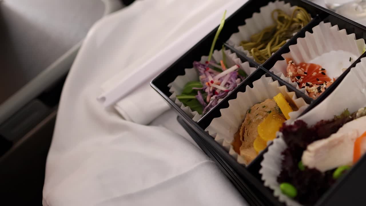 A person uses chopsticks to savor sushi and sides from a bento box on a white tablecloth.