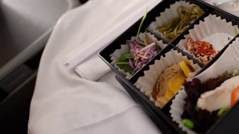 a person uses chopsticks to savor sushi and sides from a bento box on a white tablecloth.
