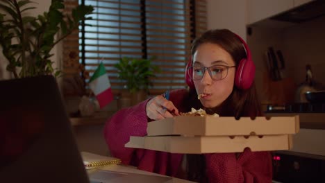 Confident-brunette-girl-in-red-wireless-headphones-and-a-sweater-studies-foreign-languages-with-the-help-of-online-lessons-takes-notes-and-eats-pizza-in-a-modern-apartment-in-the-kitchen