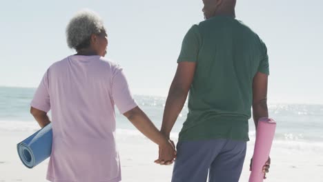 Feliz-Pareja-Afroamericana-Caminando,-Tomándose-De-La-Mano-Y-Colchonetas-De-Yoga-En-La-Playa,-En-Cámara-Lenta