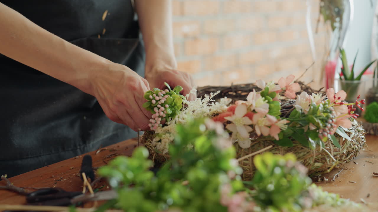 Close up of florist hands using orange garden shears to cut green stems and pink berries while creating floral wreath arrangement on wooden table with fresh leaves blossoms in natural handmade project