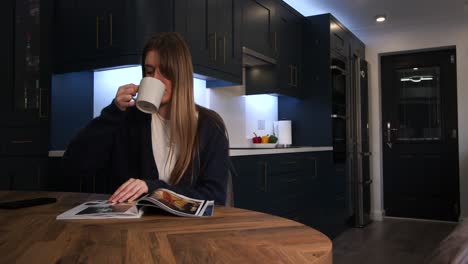 Weibliches-Model-In-Blauer-Strickjacke,-Bolero-Oberteil,-Trinkt-Aus-Einer-Tasse,-Liest-Eine-Zeitschrift-Und-Blickt-Auf-Das-Telefon-An-Einem-Küchentisch-In-Einer-Modernen-Küche