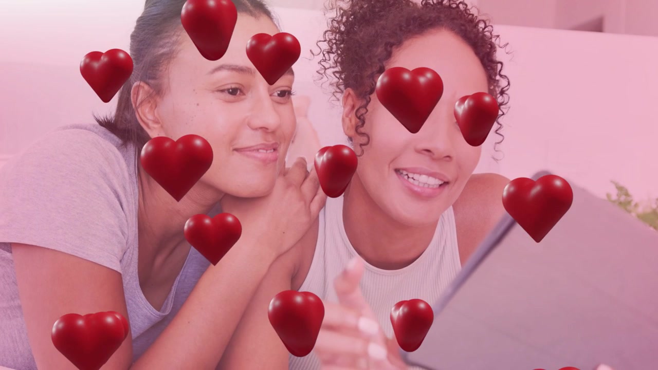 Two women reclining on pink couch pal leaning in tapping tablet sharing social post hearts floating