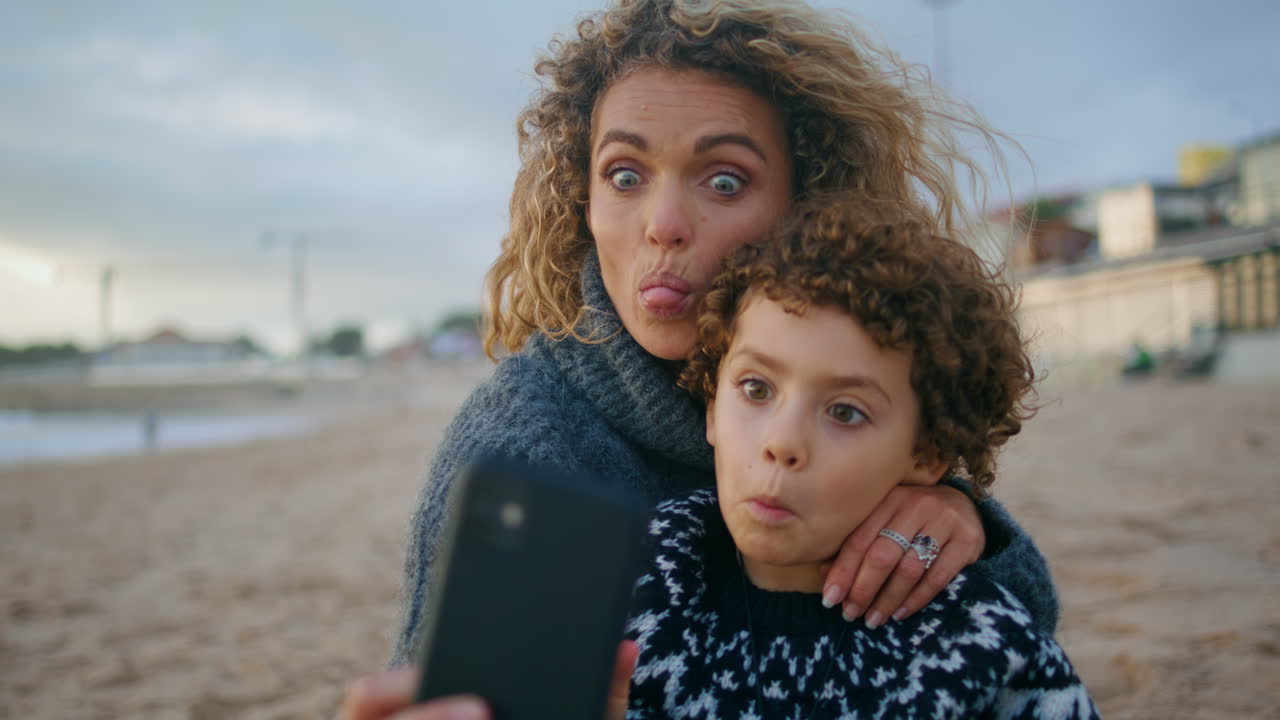 Familj tar rolig selfie på höststrand, närbild. Glad mor och son njuter tillsammans.