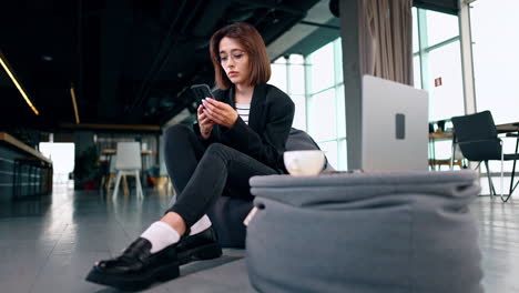 young caucasian lady sits in bean bag chair holding a phone. woman thinking over something and then types a message. low angle view.